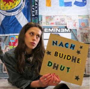 Limonead holding a handmade cardboard sign reading "Nach Buidhe Dhut" in front of a wall of posters and a Scottish flag